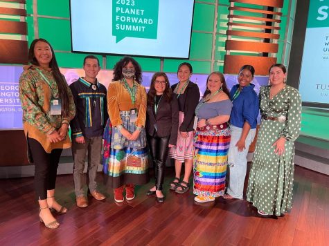 Eight members of the indigenous correspondents program of Planet Forward stand in front of a TV screen displaying the Planet Forward logo.