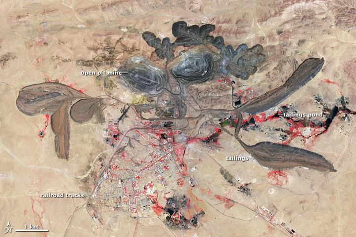 The Bayan Obo mine in 2006. The image is false-colored, showing vegetation in red, grassland in light brown, rocks in black and water surfaces in green. Two circular open-pit mines are visible, as well as a number of tailings ponds and tailings piles.