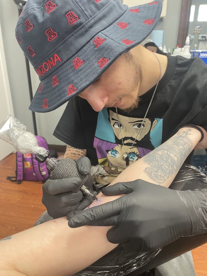 Tattoo apprentice Hunter Burns tattoos a cartoon frog from Spark Project Collective’s flash sheets on a client. 