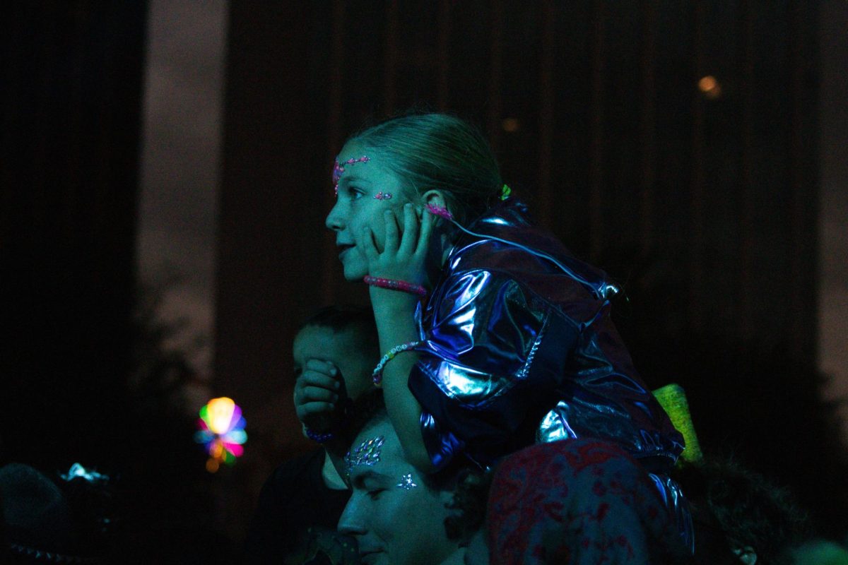 A young child with glitter and gems  sits atop her equally sparkly parent, waiting for the next act to take the stage at DUSK Music Festival on Nov. 15. at Jácome Plaza.